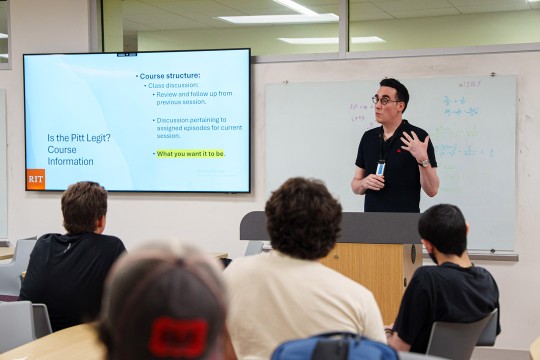 'A man in black polo stands at a podium talking to students in a classroom'