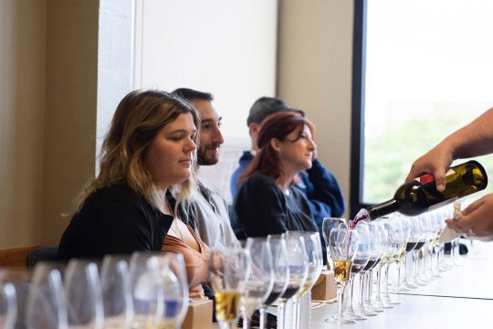 'adults sit in front of wine glasses that are being filled with wine samples in a brightly lit room'