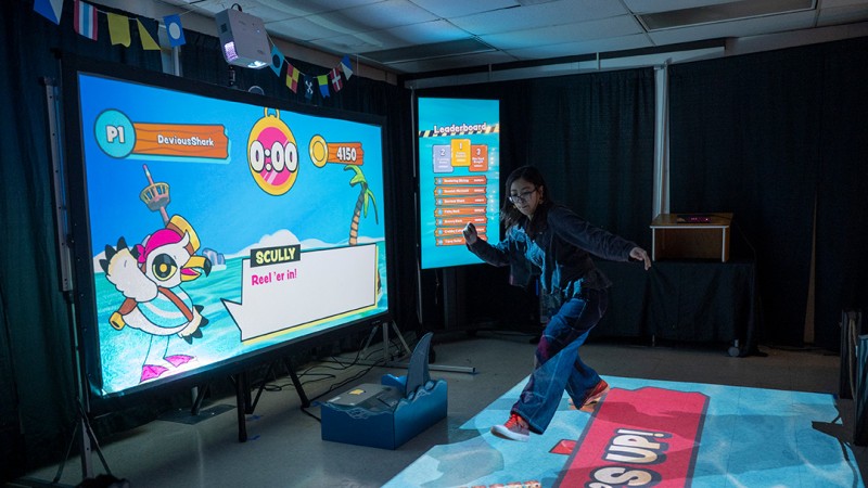 A seagull on a projected screen showing the Overboard game, with a water projected on to a floor. A student is in midair, jumping to land on wood boards to stay out of the projected water 