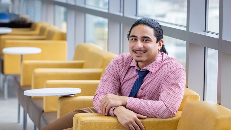 Gabe McMurtrie is wearing a pink patterned dress shirt and a navy tie and is seated in a modern lounge area with mustard-yellow armchairs and small white round tables.