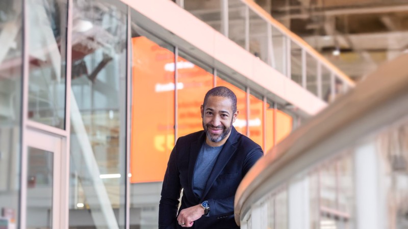RIT alumn Jason Blythe is wearing a dark blazer and gray shirt. He is leaning casually on a railing inside a modern building with glass walls and an orange digital display in the background. 