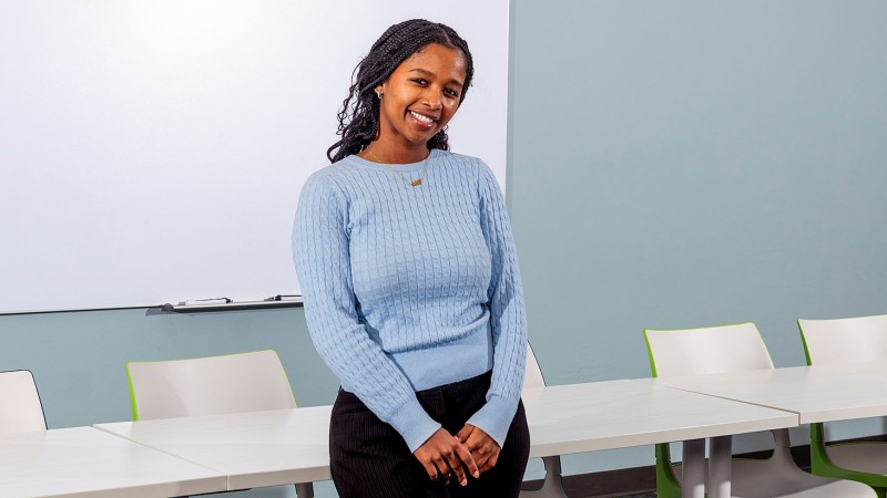 Person standing in a classroom with white tables and a whiteboard, wearing a light blue knit sweater and dark pants.