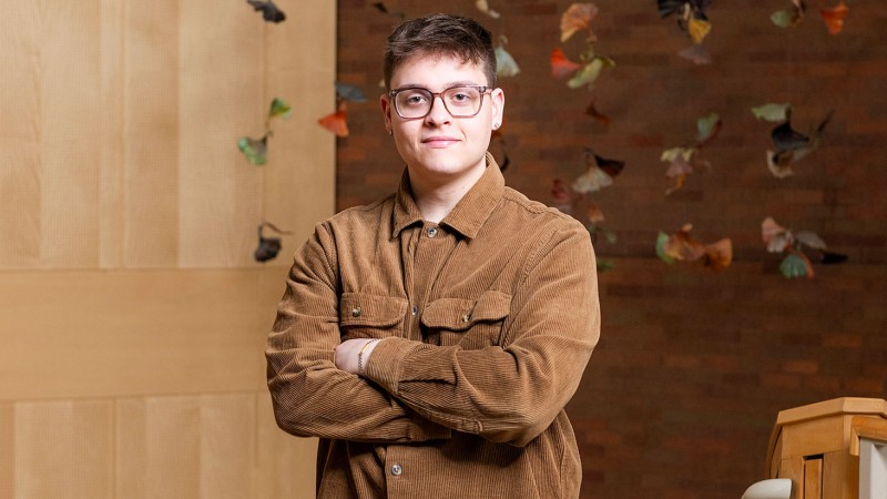 Person standing with arms crossed in front of a wooden wall decorated with hanging art pieces, wearing a brown corduroy jacket.