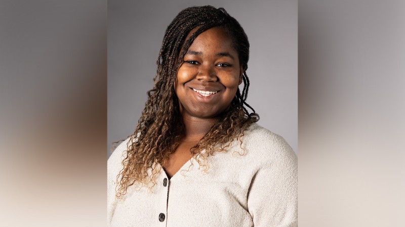Headshot of Kierstin Delk, a smiling Black woman with long braided hair wearing a cream-colored button-up cardigan against a gray background.