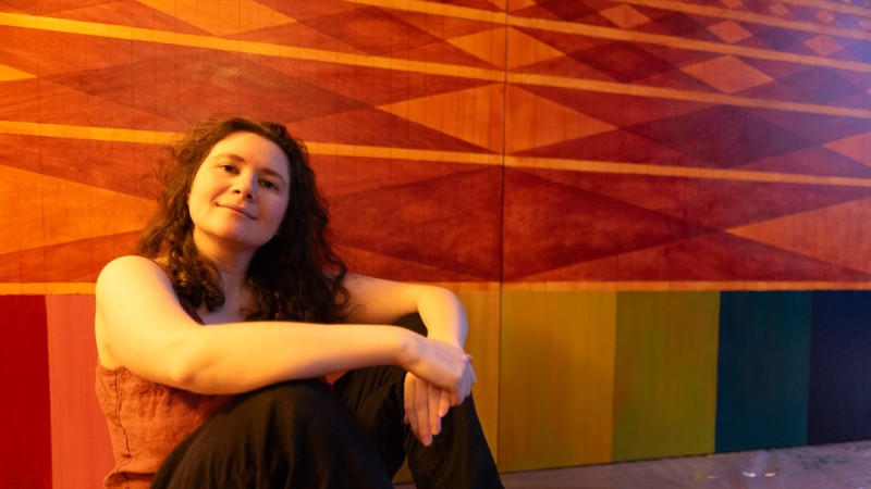 A woman with long curly brown hair sits on the floor, leaning forward with her arms crossed over her knees. She is smiling gently at the camera against a vibrant backdrop of a large wall painting featuring orange and red geometric diamond patterns above a row of multicolored vertical stripes.