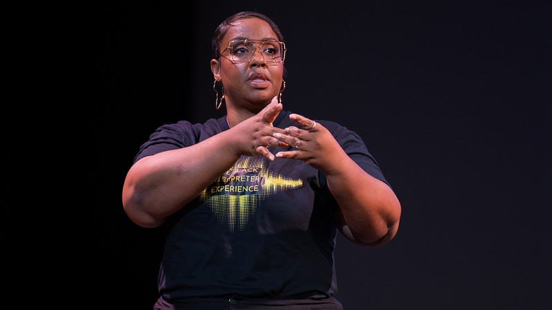 Yalanda Allgood standing on a dark stage, speaking and signing. She is wearing a black t-shirt that reads Black Interpreter Experience.