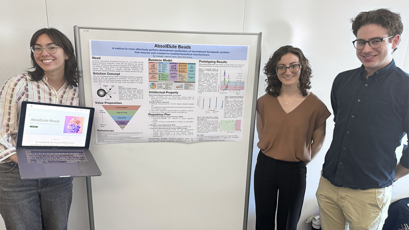 Three master's students. One standing to the left of a research poster holding a laptop. Two standing to the right of the research poster and smiling.