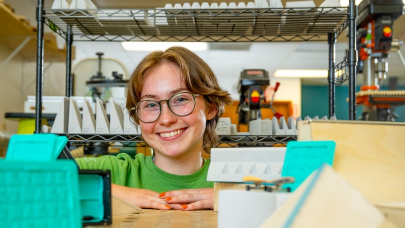 Person wearing glasses and a green shirt leans on a workbench in a makerspace or fabrication lab. In the foreground are miniature skateboarding ramps and 3D-printed objects, with shelving, tools, and workshop equipment visible in the background.