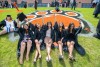 five college students wearing graduation regalia sitting on the ground in front of a tiger paw painted in the grass.