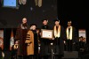 five people posing for photo wearing graduation regalia and holding a framed diploma.