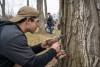 John Serafim is pictured in a blue hoodie and tan hat tapping a maple tree for sap.