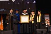 A group of people in academic regalia stand on stage, with one holding an honorary diploma