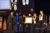 A group of people in academic regalia stand on stage, with one holding an honorary diploma