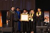 A group of people in academic regalia stand on stage, with one holding an honorary diploma