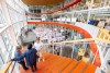 A wide camera shot of the inside of a building where students are presenting posters and people stand on stairs.