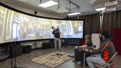 Student stands in front of super wide screen in lab displaying the inside of a mansion while other students observe.