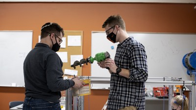 One student holding a drill and one student holding a bucket, working in a engineering lab.