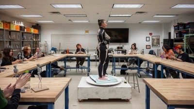 a woman wearing a skeleton bodysuit poses in the middle of a room as students make clay sculptures.