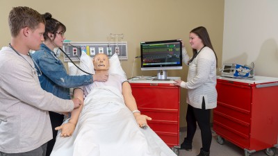 3 students using a stethoscope on a dummy patient and a monitor with data readouts