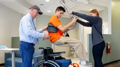 Two people assist a third middle person using a lift to transfer the middle person out of a wheelchair to a bed.