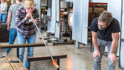 woman blows into a tube with molten glass on the end while an instructor watches.