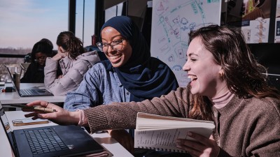 two students smile and laugh as they point to a laptop screen.