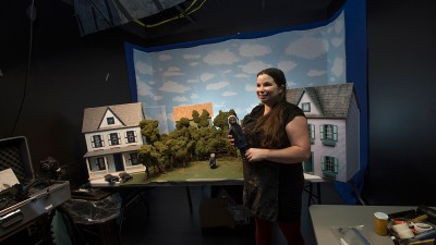 a student holds a doll in front of a scene of two models houses with a woods in between.