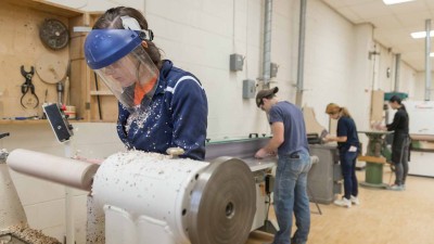 student wearing a protective mask turns wood on a lathe.