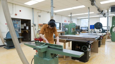 student runs a piece of wood through a jointer in a room with table saws.