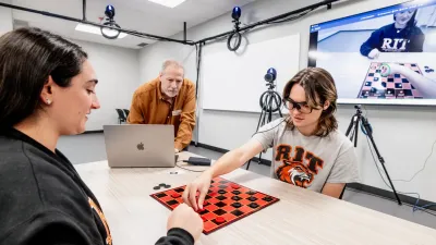 A student using an eye-tracking device plays chess with another student. The devices footage is shown on a TV in thebackground.