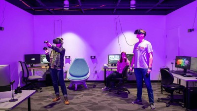 Two students stand wearing VR goggles and holding controllers in room with chairs and computers