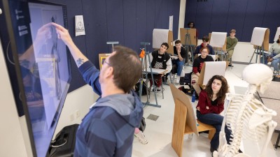 a professor gestures toward a T V screen as the class watches.