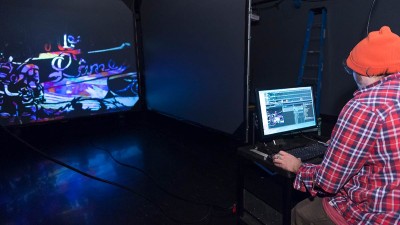 student works on a laptop as a scene is projected onto a large screen in a dark room.