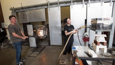 A visiting artist put glass in a kiln while a student uses a glass printer.