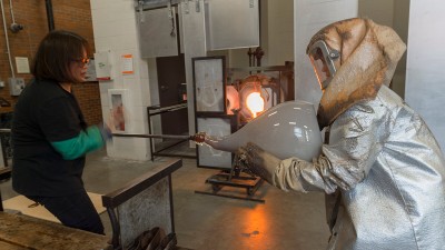 a person in a protective suit holds a large glass piece while another person connects to it with molten glass on a pole.