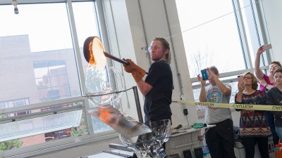 A student pours hot glass into a glass bowl as a crowd takes pictures behind him.