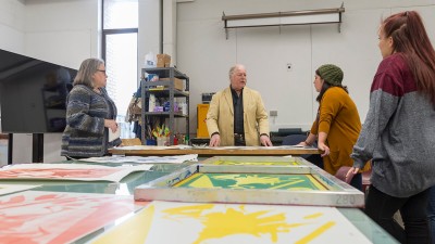 a visiting artist talks to students in front of a table with colorful prints laid out.