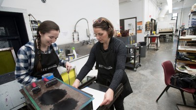 faculty member feeds a sheet of paper into a press as a student watches.