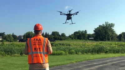 man wearing a vest identifying him as a U A V pilot flies a drone over a field.