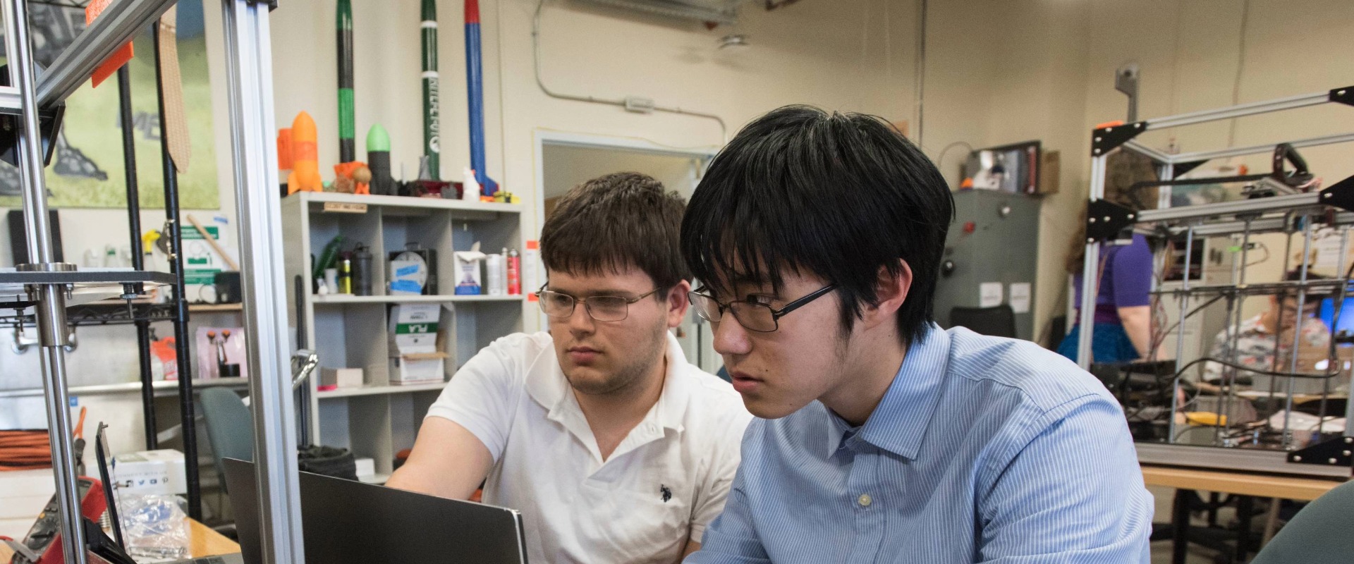 two students focus intently while looking at a computer in a room full of equipment 