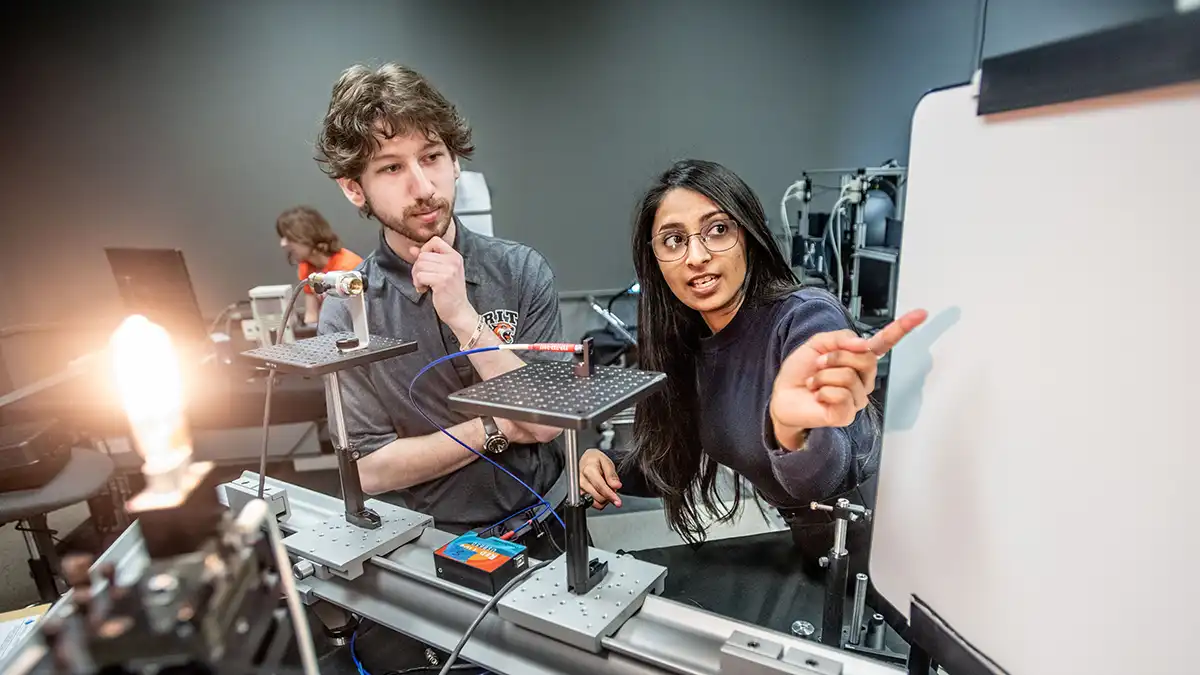 A student and instructor discuss an experiment while standing beside optical equipment and a light bulb in a lab.