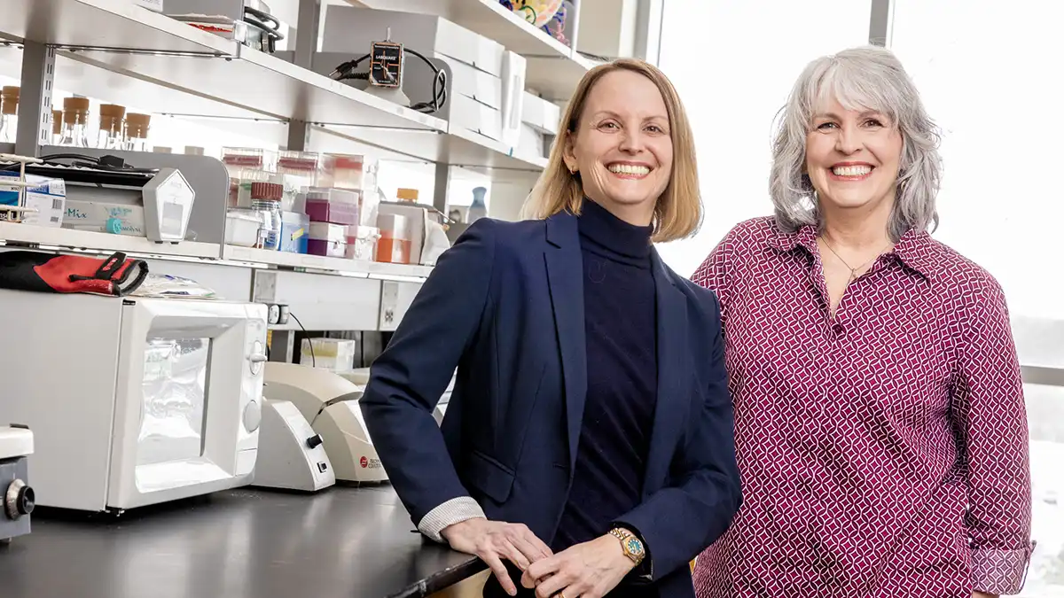 Two women stand smiling together in a laboratory with shelves of scientific supplies and equipment.