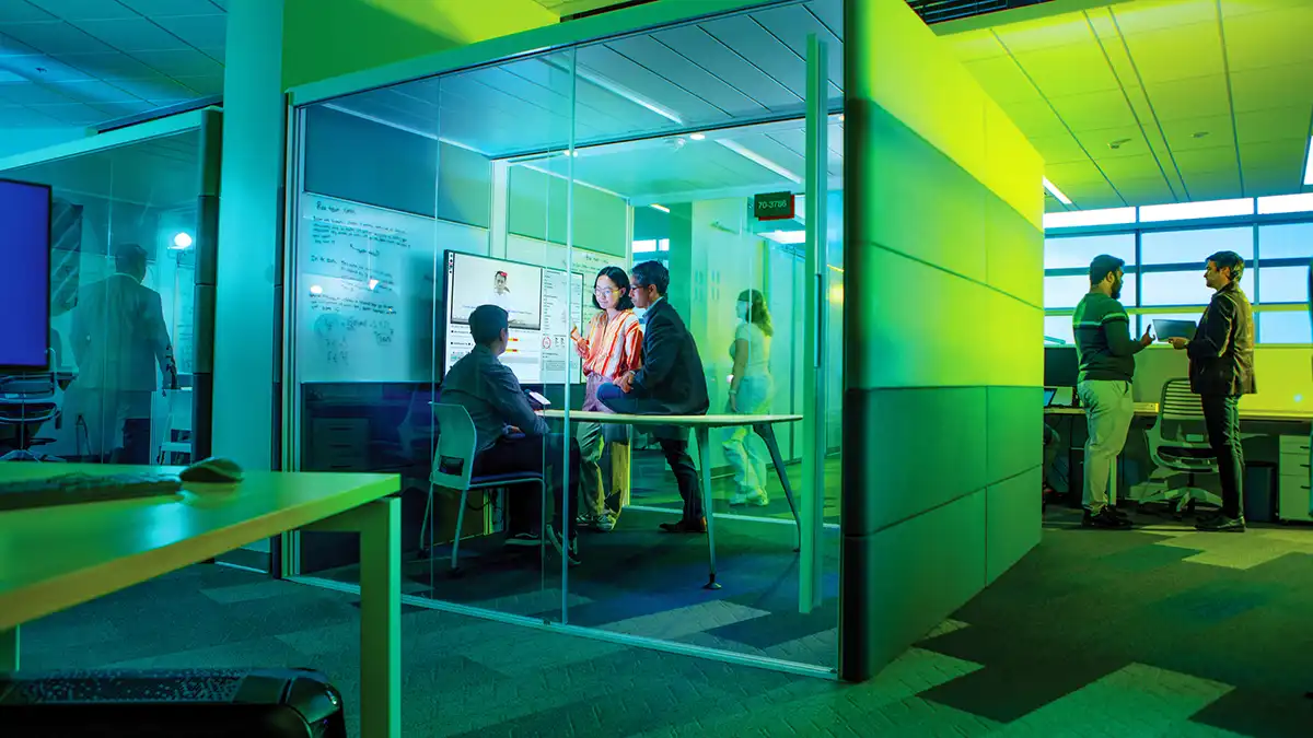 A group of people collaborate inside a glass-walled meeting room with screens and whiteboards, while others talk outside.