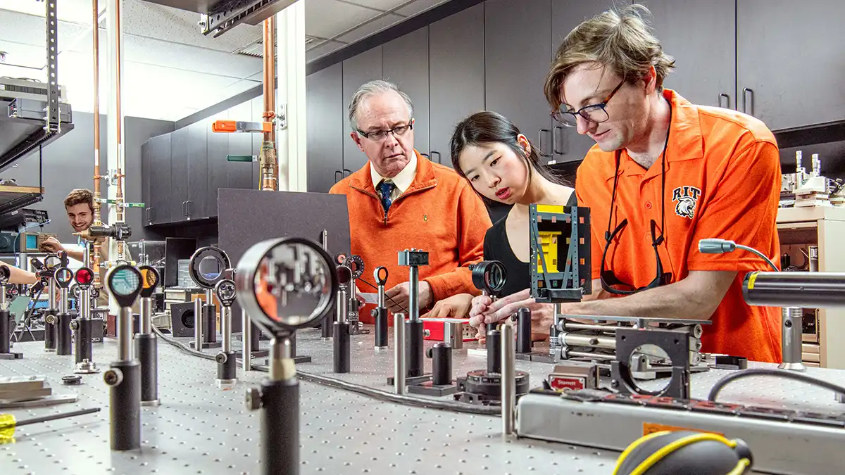Three people work together adjusting equipment on an optical physics lab bench filled with instruments and mirrors.