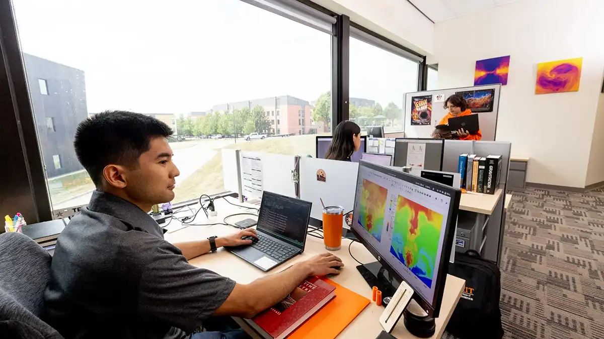 A researcher works at a computer in a bright office with multiple monitors displaying colorful scientific visualizations.