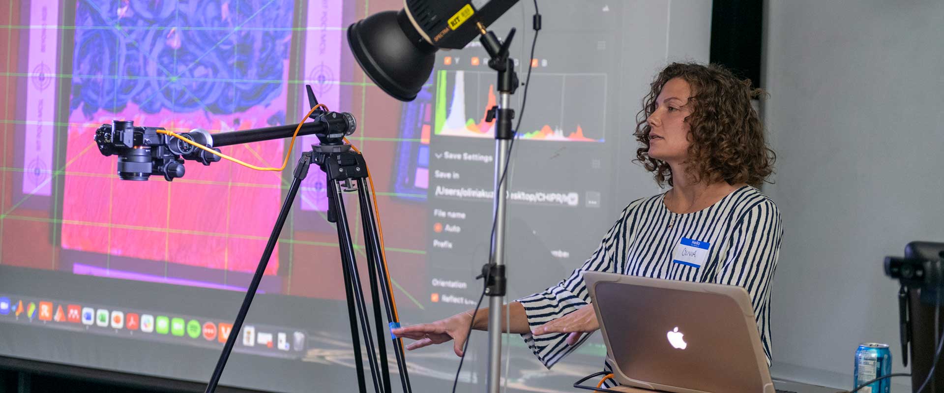 A woman gives a presentation with a laptop, camera, and projector setup, explaining digital imaging on a large screen.