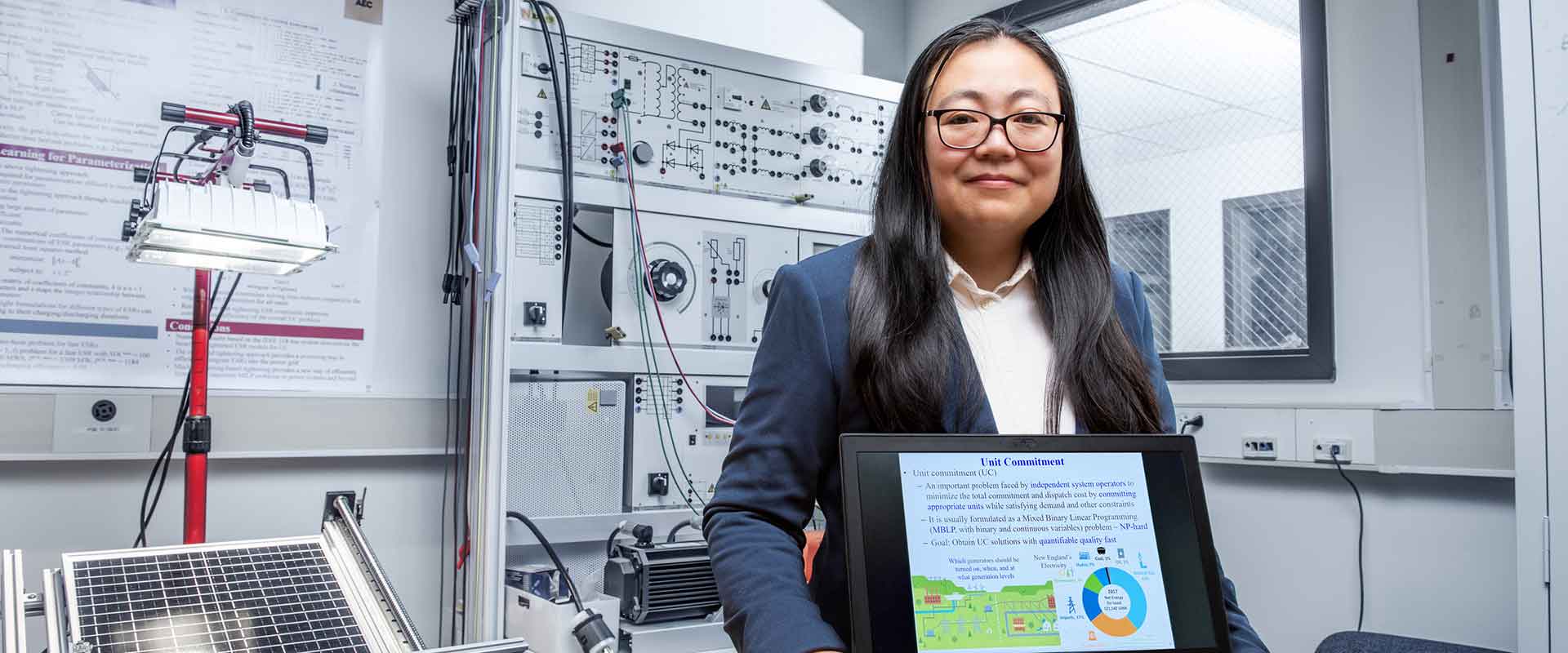 A woman stands in a lab holding a tablet displaying research slides in front of electronic equipment.