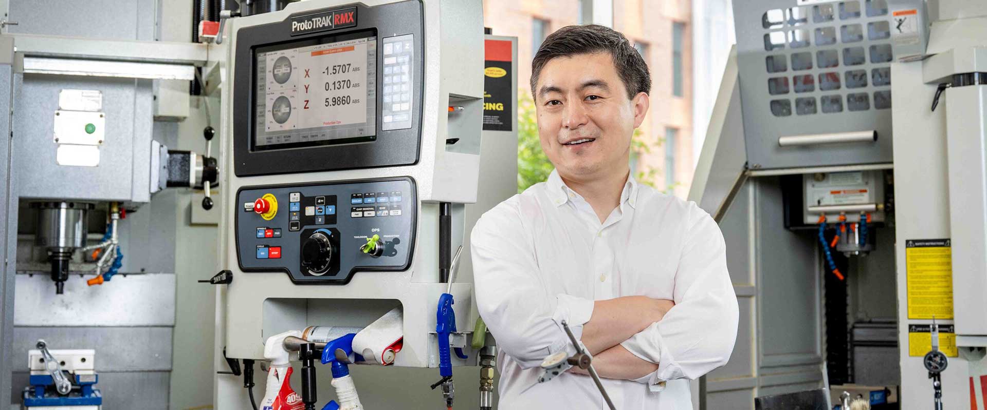 A man in a white shirt stands smiling with arms crossed in front of industrial machining equipment.