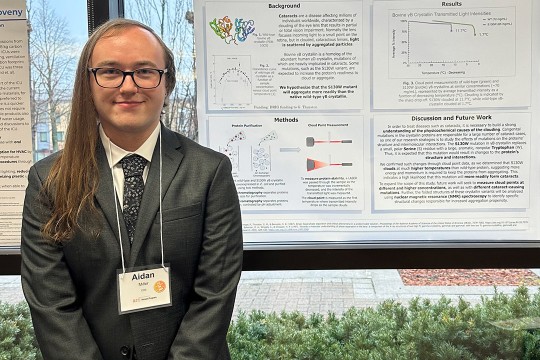 Aiden Miller is shown posing in front of their research which is hanging on a building window.