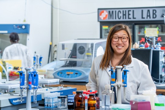 Smiling scientist in lab coat standing in a laboratory with equipment and supplies.